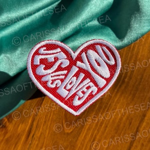 May include: Red heart-shaped embroidered patch with white lettering that says "Jesus Loves You".