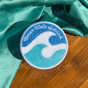 May include: A round, white embroidered patch with a blue and green wave design. The text "Ocean State of Mind" is written in white on the patch.