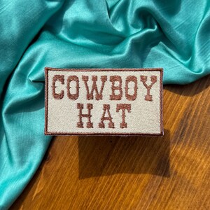 May include: A beige fabric patch with brown embroidered text that reads "COWBOY HAT" on a teal satin fabric background.