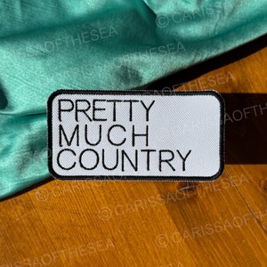 May include: White iron-on patch with black text that reads "Pretty Much Country". The patch is on a wooden surface with a teal fabric draped over the edge.