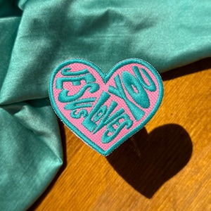 May include: Pink heart-shaped patch with the text "Jesus Loves You" in teal lettering.