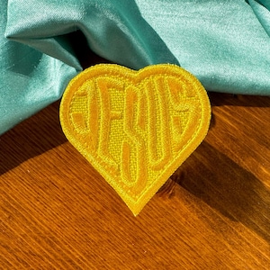 May include: A yellow heart-shaped patch with the word "JESUS" embroidered in a matching yellow thread. The patch is set against a teal fabric and a wooden surface.