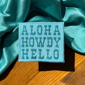 May include: A turquoise blue fabric background with a square patch that says "Aloha Howdy Hello" in a dark blue font.