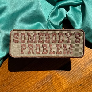 May include: A brown and green embroidered patch with the text "Somebody's Problem" in brown lettering.