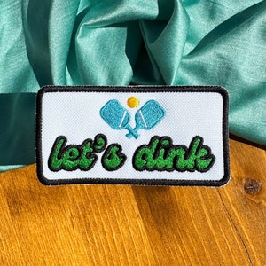 May include: A rectangular patch with a white background, black border, and the words "let's dink" in green script. Above the text are two blue pickleball paddles and a yellow ball. The patch is on a wooden surface with a teal fabric backdrop.