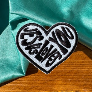 May include: White heart-shaped patch with black embroidered text that reads "Yes Love You" on a teal satin fabric background.