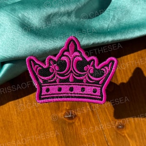 May include: A pink and black embroidered crown patch. The crown has a black background with pink details and a black outline.