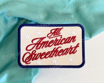 Könnte beinhalten: Ein rechteckiger, bestickter Aufnäher mit cremefarbenem Hintergrund und dunkelblauem Rand. Der Schriftzug 'All American Sweetheart' ist in roter Schreibschrift gestickt und mit kleinen weißen, auf der Fläche verteilten Sternen verziert.