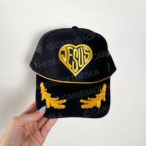 May include: Black trucker hat with a gold heart-shaped patch that says "JESUS" in a stylized font. The hat has gold embroidered details on the brim.