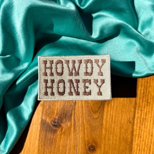 May include: A beige fabric patch with brown embroidered text that reads "Howdy Honey" on a teal satin fabric background.