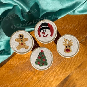 May include: Four embroidered Christmas-themed patches on a wooden surface. The patches feature a gingerbread man, a snowman, a reindeer, and a Christmas tree.