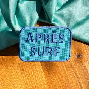 May include: A blue embroidered patch with the text "APRÈS SURF" on a wooden surface with a teal satin fabric draped over it.