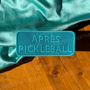 May include: A teal blue rectangular patch with the text "APRES PICKLEBALL" in white lettering. The patch is lying on a wooden surface.