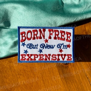 May include: A white patch with blue and red embroidered text that reads "Born Free * But Now I'm * Expensive". The text is surrounded by red stars.