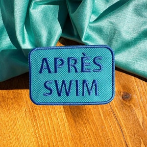 May include: A blue and white embroidered patch with the text "APRÈS SWIM" on a wooden surface with a teal satin fabric draped over it.