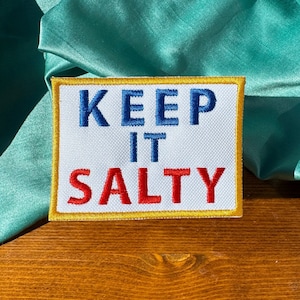May include: White embroidered patch with yellow border. The text "KEEP IT SALTY" is embroidered in blue, red, and white.
