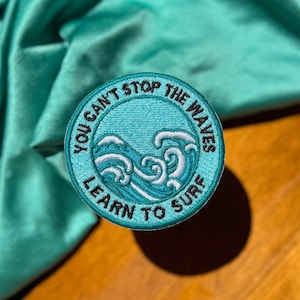 May include: A blue and white embroidered patch with the text "You can't stop the waves, learn to surf".