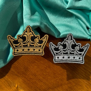 May include: Two embroidered crown patches, one gold and one silver, on a teal fabric background.