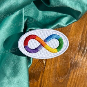 May include: White oval embroidered patch with a rainbow infinity symbol. The patch is on a wooden surface.