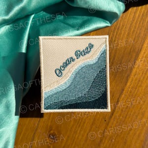 May include: A beige and blue embroidered patch with the text "Ocean Daze" in a cursive font. The patch features a stylized design of ocean waves.