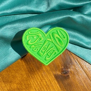 May include: Green heart-shaped patch with the word "Yes, Queen" embroidered in a 3D style. The patch is on a wooden surface with a teal satin fabric draped over it.