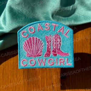 May include: A turquoise blue embroidered patch with the text "Coastal Cowgirl" in pink lettering. The patch features a pink seashell and a pair of pink cowboy boots.