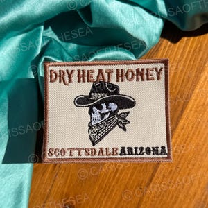 May include: A beige embroidered patch with brown stitching. The patch features a skull wearing a cowboy hat and a bandana. The text "DRY HEAT HONEY" is above the skull and "SCOTTSDALE ARIZONA" is below the skull.