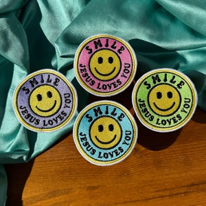May include: Four embroidered iron-on patches with a smiley face and the text "SMILE JESUS LOVES YOU" in a circle. The patches are in different colors: pink, green, light blue, and yellow.