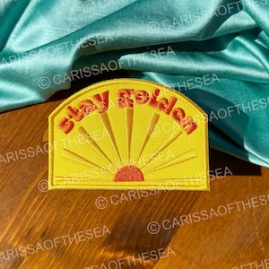 May include: Yellow embroidered patch with a sun and the text "Stay Golden" in red. The patch is on a wooden surface with a turquoise fabric draped over it.