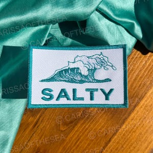 May include: A white patch with a teal border featuring a blue wave graphic and the word "SALTY" in teal lettering.