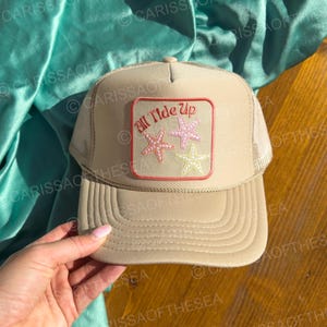 May include: A beige trucker hat with a mesh back. The hat has a red and white patch with the text "All Tide Up" and three pink and yellow starfish.