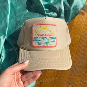 May include: A beige mesh trucker hat with a patch featuring a sun, waves, and the text "Ocean Haze".