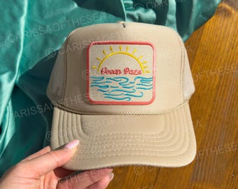 Op de afbeelding: Een beige mesh trucker cap met een patch met een zon, golven en de tekst "Ocean Haze".