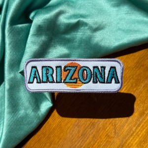 May include: White embroidered patch with the word "ARIZONA" in blue lettering and a sunset graphic in orange, yellow, and blue.