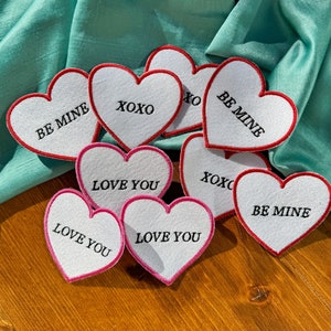 May include: Nine white felt heart-shaped patches with red and pink borders. The patches have different messages on them, including "Be Mine", "Love You", and "XOXO".