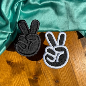 May include: Two peace sign patches, one black with white stitching and one white with black stitching, on a wooden surface.
