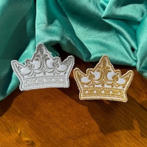 May include: Two embroidered crown patches, one silver and one gold, on a green fabric background.