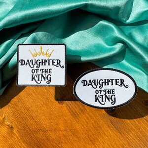 May include: Two white fabric patches with black text and a gold crown. The patches say "Daughter of the King". One patch is square and the other is oval.