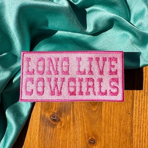 May include: Pink embroidered patch with the text "Long Live Cowgirls" on a wooden surface.