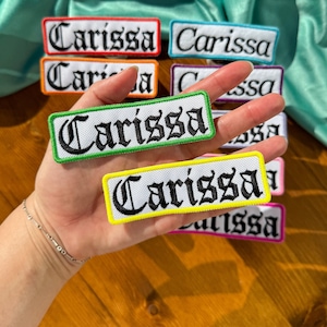May include: A collection of iron-on patches with the name "Carissa" embroidered in black on white fabric. The patches are in various colors, including red, orange, yellow, green, blue, purple, and pink.