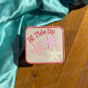 May include: A beige fabric patch with a red border and the text "All Tide Up" in pink lettering. Three pink and white starfish are embroidered on the patch.