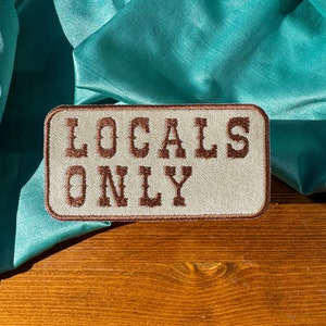 May include: A beige embroidered patch with brown stitching that reads "LOCALS ONLY".