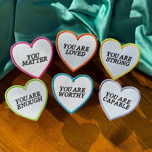 May include: Six white felt heart-shaped patches with colorful borders. Each patch has a different positive affirmation embroidered in black: "You Are Loved", "You Are Strong", "You Are Enough", "You Are Worthy", "You Are Capable", and "You Matter".