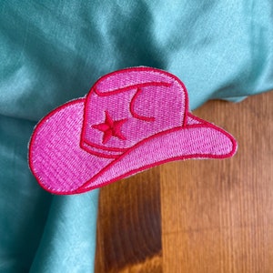 May include: Pink felt cowboy hat with a white star embroidered on the front. The hat is on a wooden surface.