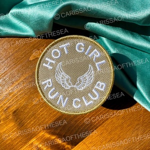 May include: A round, beige patch with white embroidery. The patch says "Hot Girl Run Club" with a pair of white angel wings in the center.