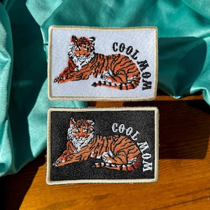 May include: Two embroidered iron-on patches with a tiger design and the text "Cool Mom". One patch is white with black text and the other is black with white text.