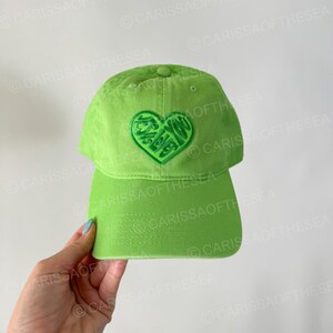 May include: A bright green baseball cap with a green heart-shaped logo embroidered on the front. The logo has a stylized design with the word "LOVE" inside the heart.