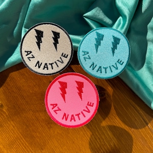May include: Three embroidered patches with a smiley face design. Each patch features a lightning bolt design and the text "AZ NATIVE". The patches are in white, turquoise, and pink.