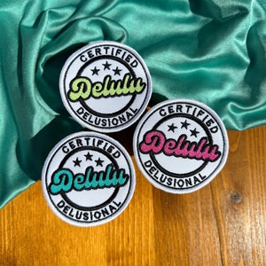May include: Three white round embroidered patches with the text "CERTIFIED DELUSIONAL" and three stars. The patches have different colored text: green, pink, and teal.
