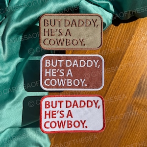 May include: Three embroidered patches with the text "But Daddy, He's a Cowboy." The patches are in different colors: beige, brown, and white with red stitching.